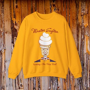May include: A mustard yellow crewneck sweatshirt featuring a graphic of a Mister Softee ice cream cone character with a smiling face, wearing a bowtie. The text "Mister Softee" appears above the graphic, and "Serving The Very Best" is printed below.
