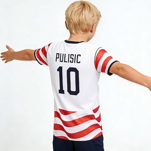 Pode incluir: Camisola de futebol branca com detalhes em vermelho e azul, com o nome "PULISIC" e o número "10" em azul marinho. A camisola tem detalhes de riscas vermelhas e brancas nas mangas e na bainha. A pessoa está a usar calções azuis marinho.