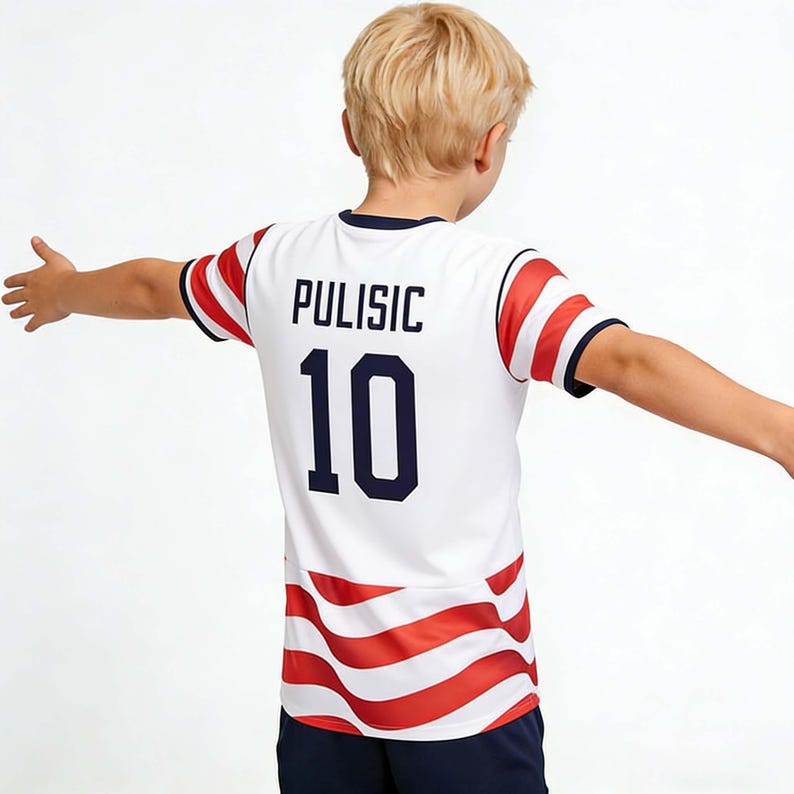 May include: White soccer jersey with red and blue accents, featuring the name "PULISIC" and the number "10" in navy blue. The jersey has red and white striped details on the sleeves and lower hem. The person is wearing navy blue shorts.
