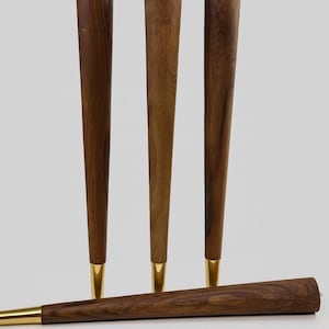 May include: Four wooden furniture legs with a tapered design. The legs are a warm brown color with a gold-colored metal tip. Three legs stand upright, while the fourth lies horizontally. The legs are designed for furniture.