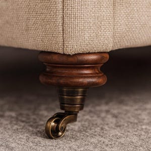 May include: Close-up of a beige upholstered furniture piece with a dark wood base and antique brass caster wheel. The fabric has a textured appearance. The caster wheel is a decorative detail.