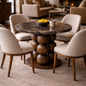 May include: A round dining table with a dark marble top and a stacked wooden base. Four beige upholstered chairs with wooden legs surround the table. A bowl of lemons and a mug sit on the table, creating a warm, inviting dining area.