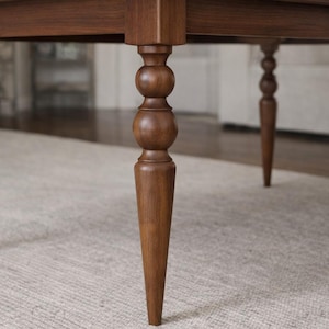 May include: Close-up of a wooden table with four legs. The legs are a dark brown color and have a turned design with a bulbous shape near the top. The table legs are tapered and rest on a light-colored rug.