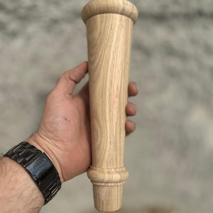 May include: A light-colored, unfinished wooden furniture leg with a turned design. The leg has a rounded top and a decorative base, showcasing the natural wood grain. The leg is held in a hand, suggesting its size and form.