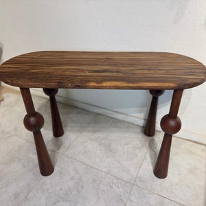 May include: A dark brown wooden table with an oval top and four legs. The legs are a dark brown colour with a ball-shaped accent in the middle. The table is approximately 122 cm long and 61 cm wide.