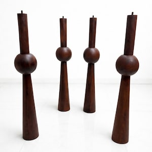 May include: Four dark brown wooden furniture legs. Each leg features a cylindrical top, a spherical mid-section, and a conical base. The legs are positioned on a white tiled floor, evoking a minimalist or mid-century modern style.