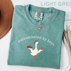 May include: Light green t-shirt with the text "outnumbered by boys" and a cartoon goose wearing a baseball cap and holding a football and a red truck. A silver necklace is draped across the shirt.