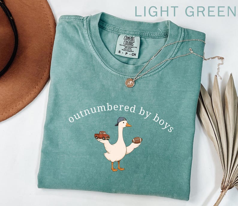 May include: Light green t-shirt with the text "outnumbered by boys" and a cartoon goose wearing a baseball cap and holding a football and a red truck. A silver necklace is draped across the shirt.