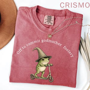 Könnte beinhalten: Karmesinrotes T-Shirt mit einer Grafik einer Froschfigur mit Hexenhut, die einen Roller fährt. Der Text "Off to commit godmother foolery" ist darüber angeordnet. Eine silberne Halskette mit Anhänger liegt auf dem Shirt.