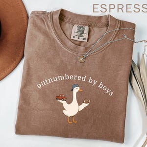 May include: Espresso-colored t-shirt with the text "outnumbered by boys" and a cartoon goose wearing a baseball cap, holding a toy truck and a football. The shirt has a crew neck and short sleeves. A silver necklace is draped across the shirt.