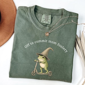 Op de afbeelding: Saliegroen t-shirt met de tekst "Off to commit mom foolery" en een afbeelding van een kikker met een tovenaars hoed op een scooter. Een gouden ketting is over het shirt gedrapeerd. Een bruine hoed staat in de linkerbovenhoek.