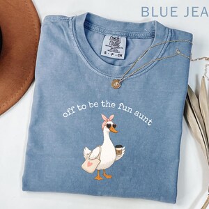 May include: A blue jean coloured t-shirt featuring a graphic of a white goose wearing a pink headband and heart-shaped sunglasses. The goose carries a tote bag and a coffee cup. The text above the goose reads, off to be the fun aunt.