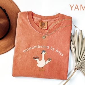 May include: A coral-colored, crew neck t-shirt with the phrase "outnumbered by boys" in white script. The shirt features a cartoon goose wearing a baseball cap and holding a football and a red truck. A gold necklace is draped across the shirt.