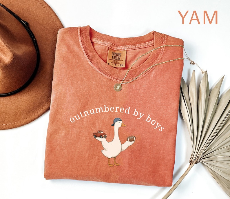 May include: A coral-colored, crew neck t-shirt with the phrase "outnumbered by boys" in white script. The shirt features a cartoon goose wearing a baseball cap and holding a football and a red truck. A gold necklace is draped across the shirt.