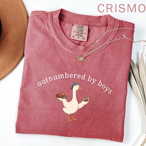 May include: Crimson-colored t-shirt with the text "outnumbered by boys" and a cartoon goose wearing a baseball cap, holding a toy truck and a football. The shirt has a crew neck and a small tag. A silver necklace with a pendant is on the shirt.