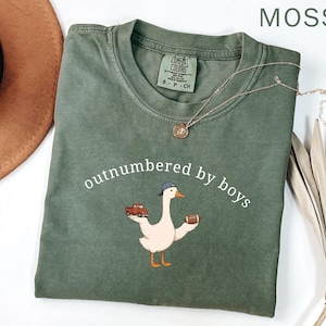 May include: Moss green t-shirt with the phrase "outnumbered by boys" and a cartoon goose wearing a baseball cap, holding a football and a red truck. A gold necklace is draped across the shirt.