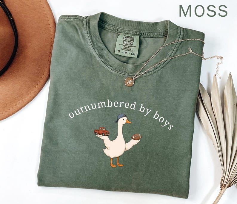 May include: Moss green t-shirt with the phrase "outnumbered by boys" and a cartoon goose wearing a baseball cap, holding a football and a red truck. A gold necklace is draped across the shirt.
