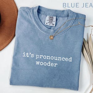 May include: A blue jean Comfort Colors t-shirt with the text "it's pronounced wooder" in white. A gold necklace with a pendant is draped across the shirt. A brown hat is visible in the upper left corner.