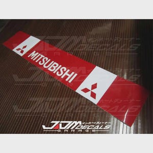 May include: A red and white Mitsubishi decal. The decal features the Mitsubishi logo and the brand name in white lettering. The rectangular decal has a glossy finish.