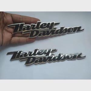 Puede incluir: Dos emblemas de Harley-Davidson. Los emblemas son plateados con letras negras. La escritura de Harley-Davidson está en una fuente estilizada. Los emblemas se muestran sobre un fondo gris claro.