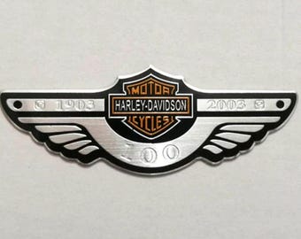 Peut inclure: Un emblème Harley-Davidson argenté et noir avec un design d'aile. L'emblème comporte le texte "MOTOR HARLEY-DAVIDSON CYCLES" et les années "1903" et "2003". L'emblème a une forme de bouclier au centre.