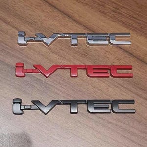 May include: Three metallic i-VTEC emblems in silver, red, and black, arranged on a wooden surface. The emblems feature a stylized font, with the "i" and "TEC" elements clearly defined. The silver emblem is at the top, followed by a red one, and a black one at the bottom.