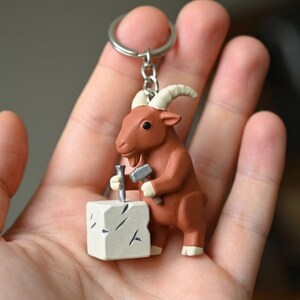 May include: A brown goat figurine keyring with white horns, holding a hammer and chisel, working on a white block. The goat is attached to a silver keyring. A unique accessory.