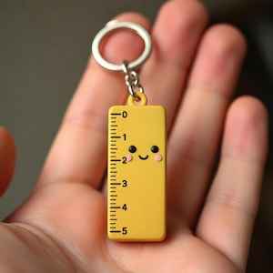 Op de afbeelding: Een gele liniaalvormige sleutelhanger met een lachend gezicht en zwarte markeringen die metingen van 0 tot 5 inch (ongeveer 12,7 cm) aangeven. De sleutelhanger heeft een zilveren ring aan de bovenkant.