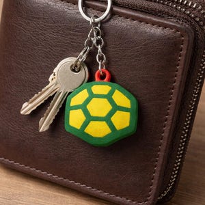 May include: A brown leather wallet with a silver zip and a keychain. The keychain features a green, yellow, and red turtle design. Two keys are attached. The wallet rests on a wooden surface.