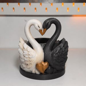 以下が含まれることがあります： 金色のハートが付いた、白と黒の白鳥の装飾品。 白鳥の首はハート型を形成しています。 白い白鳥はクリーム色の体をしており、黒い白鳥は質感があります。 ベースは黒です。