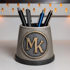 May include: A gray pen holder with a black base and a gold-colored MK emblem. The pen holder contains several pens in black, gray, and blue. The pen holder is made of a textured material.
