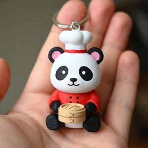 Puede incluir: Un llavero de panda chef. El panda es blanco con ojos, orejas y pies negros. Lleva una chaqueta y un gorro de chef rojos y sostiene un pequeño vaporizador de bambú. El llavero está hecho de un material duradero.