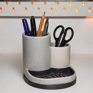 Può includere: Un organizer da scrivania grigio e bianco con due scomparti cilindrici e un piccolo vassoio. L'organizer contiene penne, matite, forbici e graffette. La base è nera.