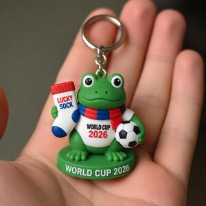 Puede incluir: Un llavero de rana verde con una camiseta blanca con el texto WORLD CUP 2026 en negro y rojo. La rana lleva una bufanda roja y azul, sostiene un calcetín blanco con el texto LUCKY SOCK en rojo y azul, y lleva un balón de fútbol.