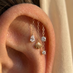 May include: Close-up of an ear with three gold chain earrings. One earring has a pear-shaped clear stone, another a small gold seashell, and the third has two clear, round stones. The earrings are delicate and elegant.