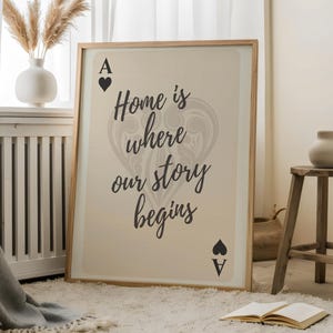 Op de afbeelding: Een ingelijste print met de tekst "Home is where our story begins" in een stijlvol lettertype. De print toont de symbolen van de schoppen aas kaart. Het kunstwerk wordt tentoongesteld op een wit tapijt, bij een raam en een houten kruk.