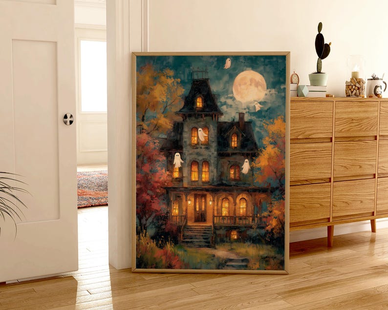 May include: A rectangular wall art print in a light wood frame depicting a multi-story Victorian house at night. The scene features a large full moon, glowing yellow windows, and several white ghost figures floating around the dark blue exterior.