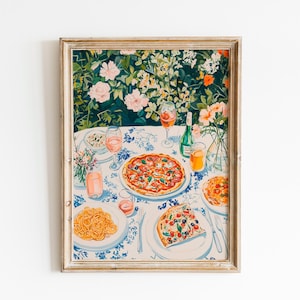 Op de afbeelding: Een ingelijste print van een tafel gedekt voor een maaltijd, met pizza, pasta, wijn en bloemen. De tafel is bedekt met een blauw-witte bloemen tafelkleed. De achtergrond toont een weelderige tuin met roze en oranje bloemen.