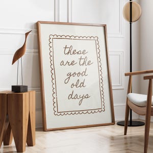 May include: A rectangular framed wall art piece featuring a light-colored background with a brown scalloped border and the brown handwritten text "these are the good old days" centered within the frame, displayed in a wooden frame.