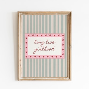May include: A framed wall art print featuring a light blue and beige vertical striped background. A central white rectangular panel contains the dark red cursive text long live girlhood, surrounded by a pink border decorated with small red hearts.