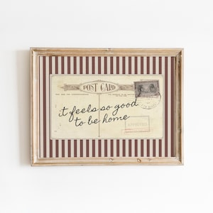 May include: Framed vintage postcard print with the text "it feels so good to be home." The postcard is set against a striped background in shades of brown and cream, encased in a wooden frame.