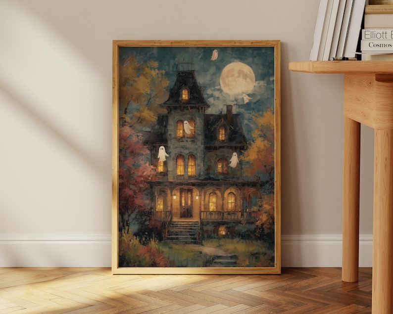 May include: A framed rectangular wall art print depicting a multi-story dark house at night with glowing yellow windows, a large full moon in the sky, and several small white ghost figures floating around the exterior of the building.