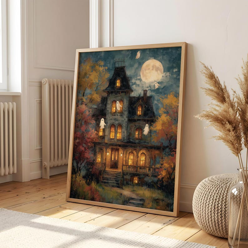 May include: A framed wall art print depicting a multi-story Victorian house at night with glowing yellow windows, surrounded by autumn trees with orange and red foliage, a large full moon in the dark blue sky, and several small white ghosts.