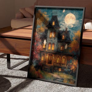 May include: A framed rectangular wall art print featuring a multi-story house with glowing yellow windows, surrounded by autumn trees in shades of orange and red. A large full moon shines in the dark blue sky, with several white ghost figures.
