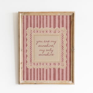 May include: A framed wall art print featuring a dusty rose and white vertical striped background with a damask pattern. Centered is a beige square bordered by cream-colored lace, containing the brown text: you are my sunshine, my only sunshine.