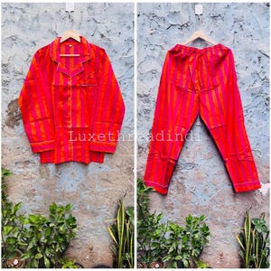 Puede incluir: Un conjunto de pijama de dos piezas con rayas verticales rojas y naranjas vibrantes. La camisa abotonada de manga larga y los pantalones a juego se muestran en perchas. El conjunto presenta ribetes blancos y el texto "Luxethreadindi".
