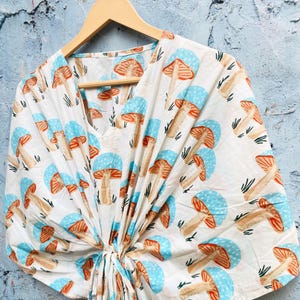 May include: A white, V-neck top with a repeating pattern of mushrooms in shades of blue, orange, and brown. The top is tied at the waist and hangs from a wooden hanger against a textured blue wall. The mushroom design is a whimsical, nature-inspired print.