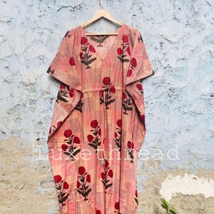 May include: A pink and red floral print kaftan dress with a V-neck and short sleeves. The dress is hanging on a wooden hanger against a textured wall. The text "Luxe thread" is visible on the dress.
