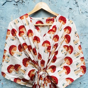 May include: White V-neck top with a repeating pattern of red and tan mushrooms. The fabric is gathered at the waist and hangs from a wooden hanger against a blue textured wall. The mushroom design is a whimsical, nature-inspired print.