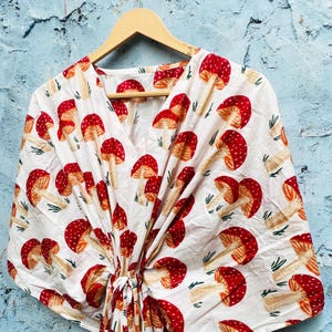 May include: White, V-neck top with a repeating pattern of red and white mushrooms. The mushrooms have red caps with white spots and beige stems. The top is hanging on a wooden hanger against a blue wall.
